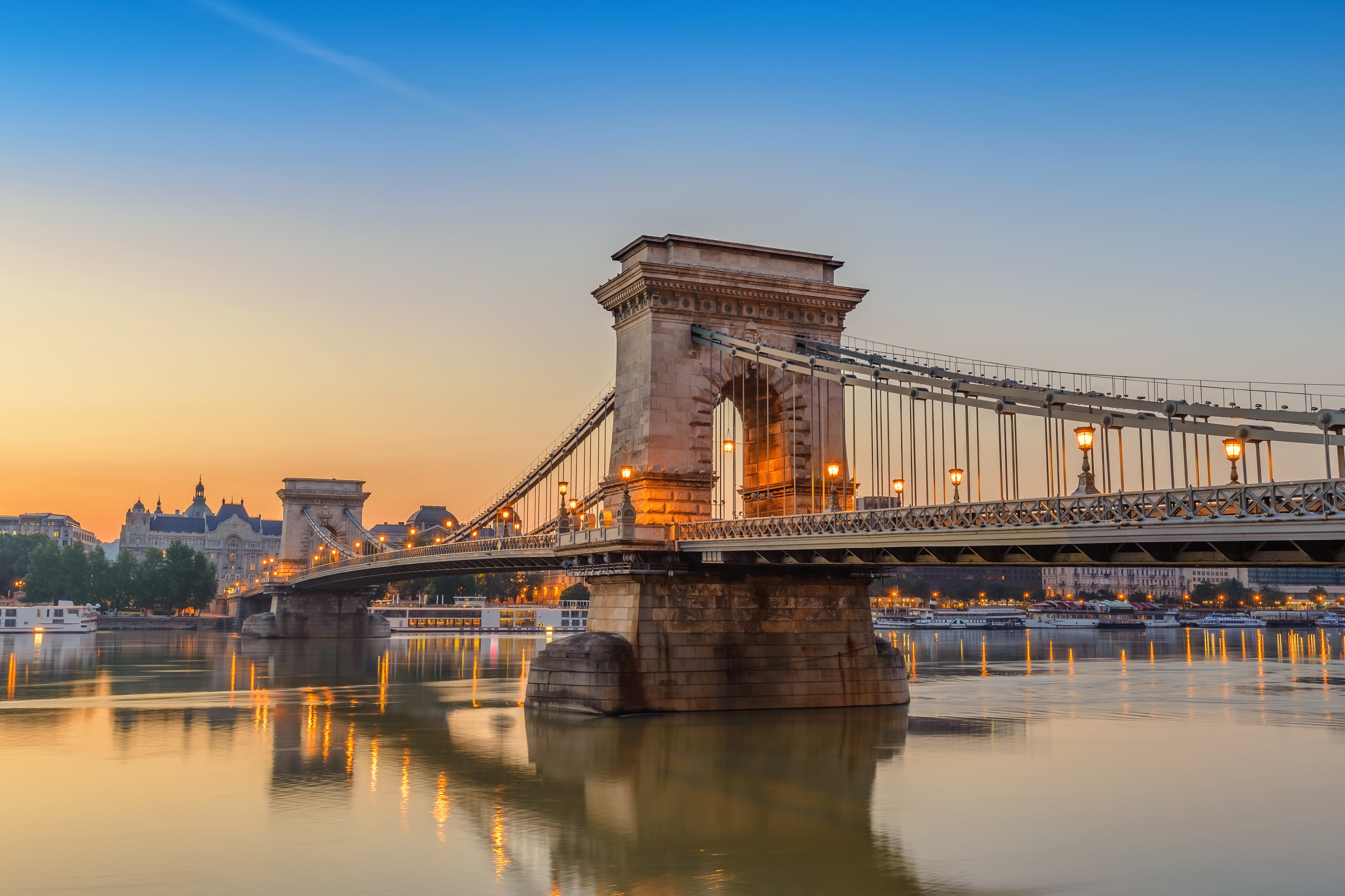 Ketjusilta Budapestissa auringonlaskun aikaan heijastuu Tonavaan, taustallaan kaupungin rakennuksia