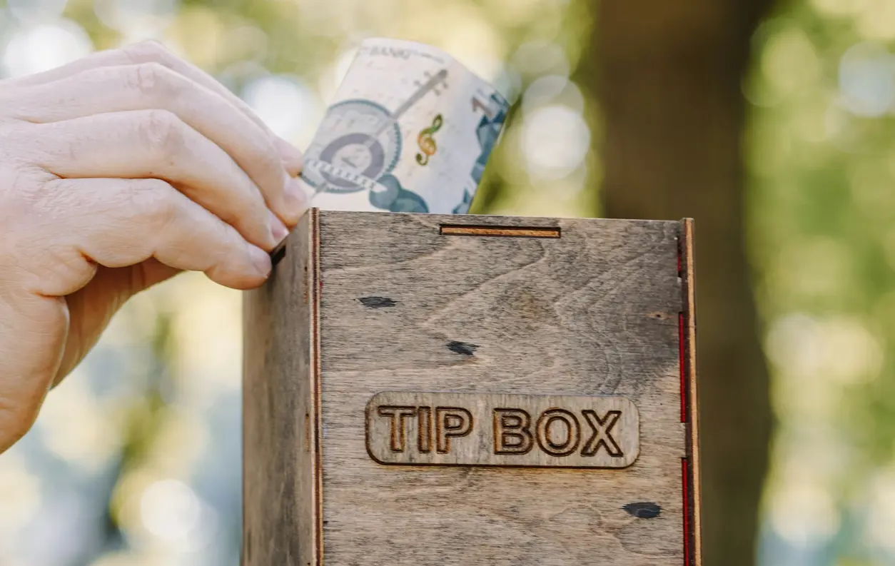 Hand lägger en sedel i en träbox märkt “Tip Box”, avsedd för dricks.