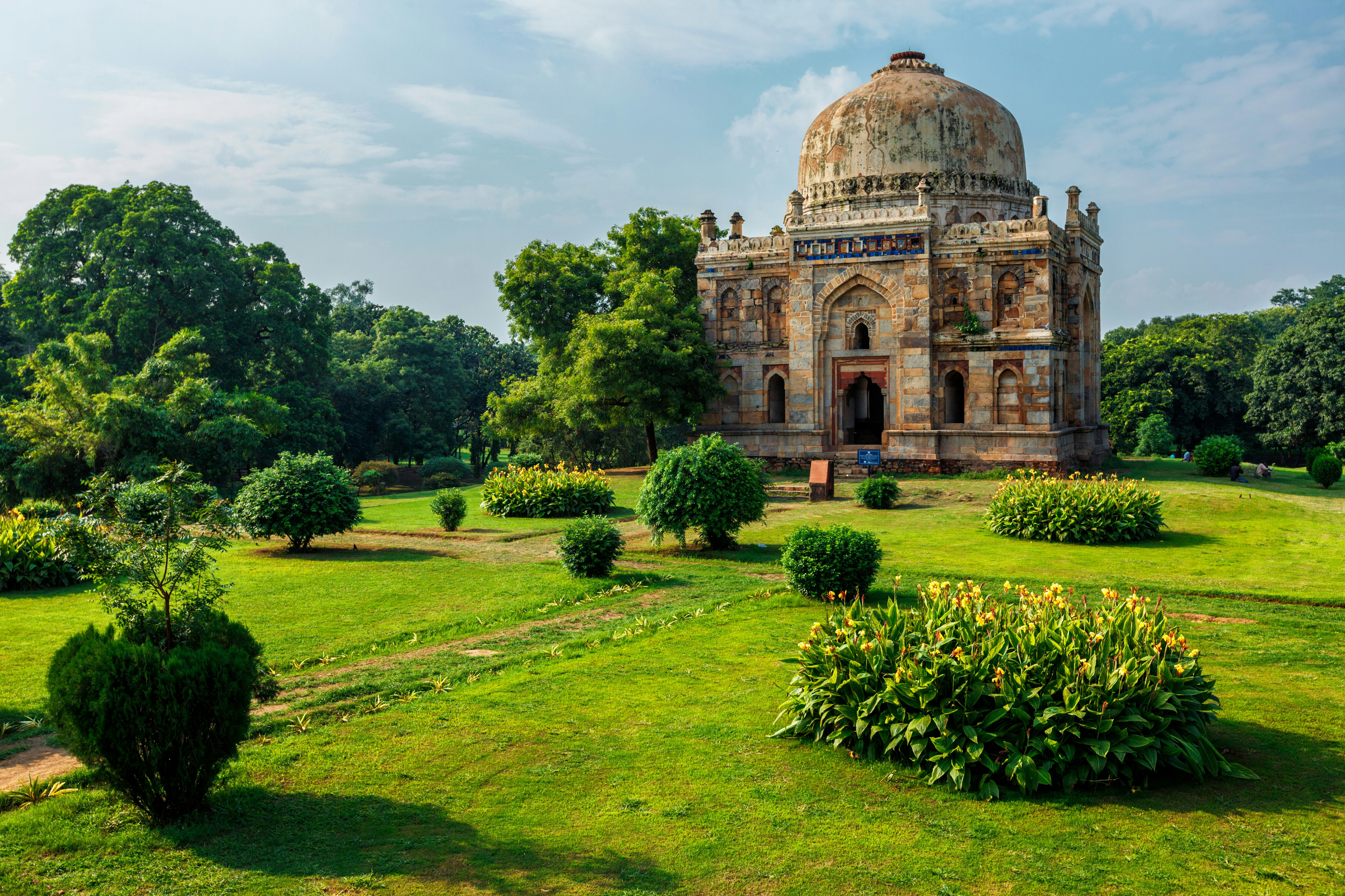 Lummig trädgård och historisk kupolformad byggnad under klarblå himmel i Delhi.
