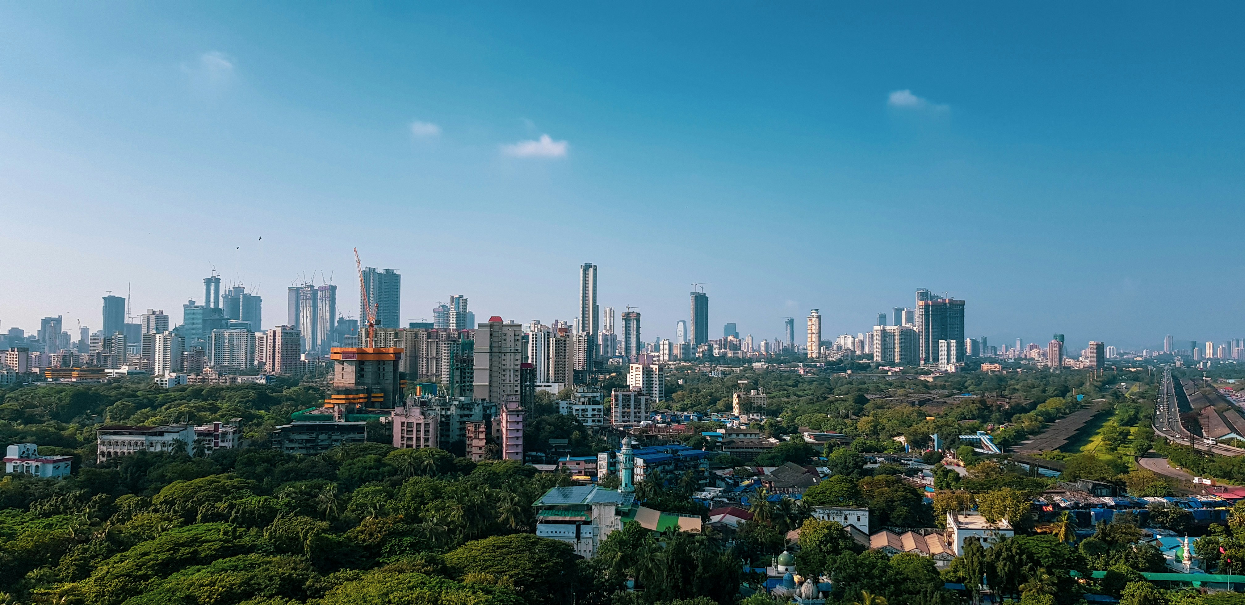 Matkusta Mumbai-Bombayhin - Panoraamanäkymä Mumbaihin, jossa on pilvenpiirtäjiä, vihreitä puistoalueita ja kirkas sininen taivas.