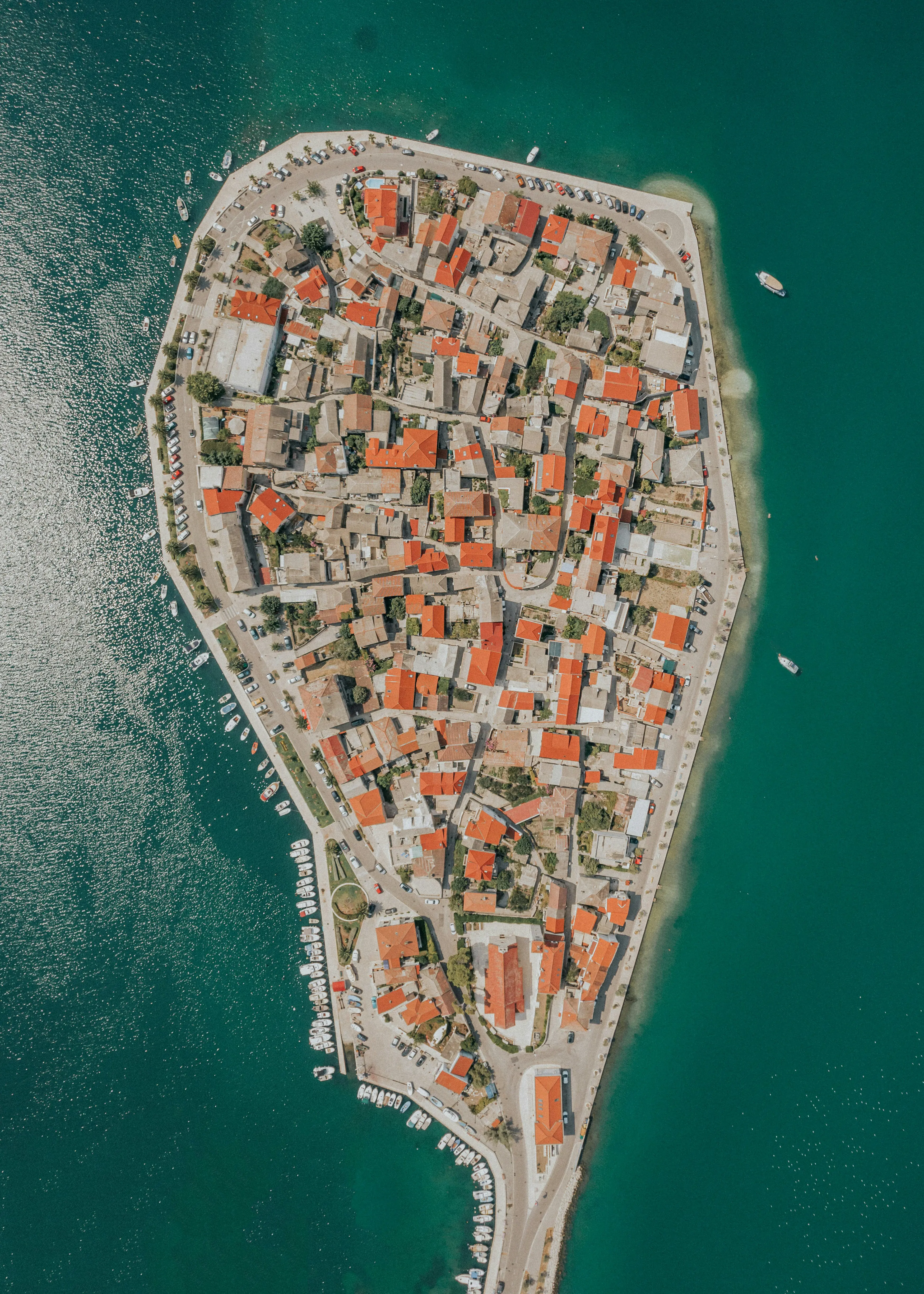 Aerial view of a European coastal town with red-roofed buildings surrounded by turquoise water and docked boats