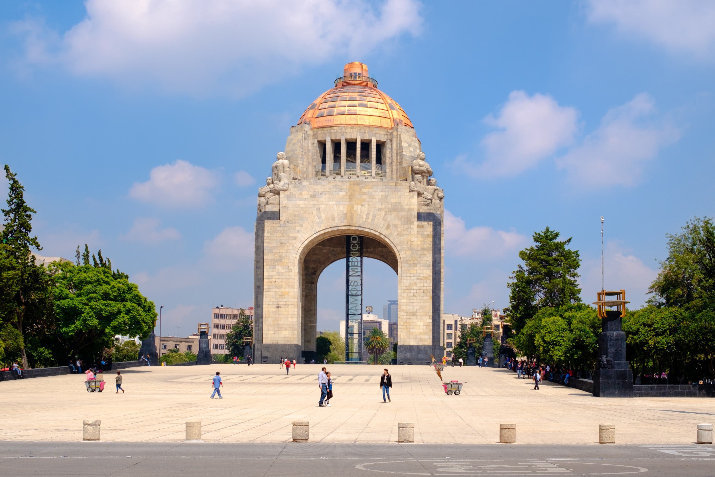 En torg som leder upp till monument i Mexico City med gröna träd omkring och blå himmel i bakgrunden