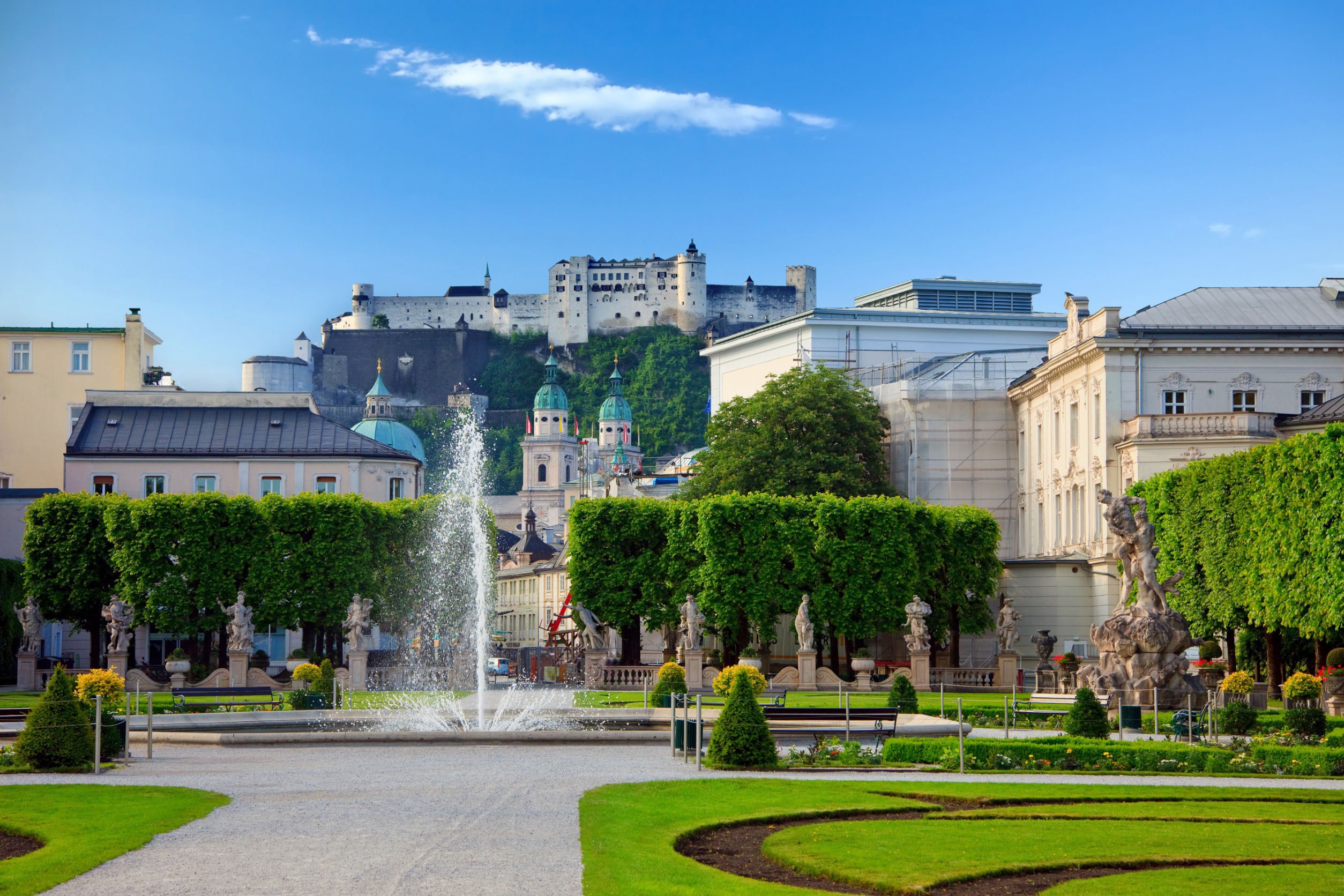 Kaunis vihreä puisto, jossa on perinteisiä eurooppalaisia rakennuksia ja suihkulähde sinistä taivasta vasten Salzburgissa