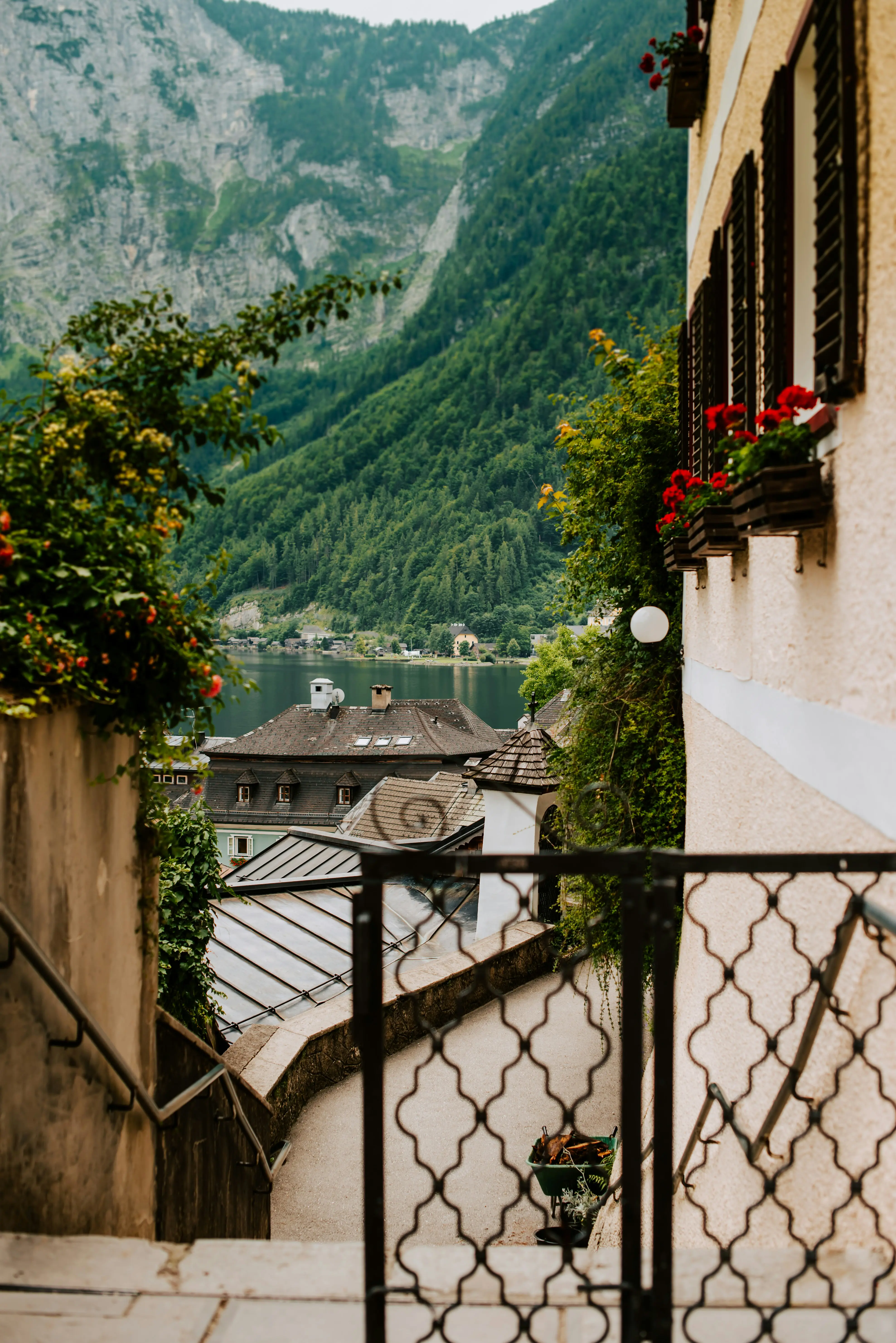 Scenic view of a European village street with ornate railing, overlooking rooftops and a lush green mountain landscape. Red flowers adorn a yellow house, adding vibrant color