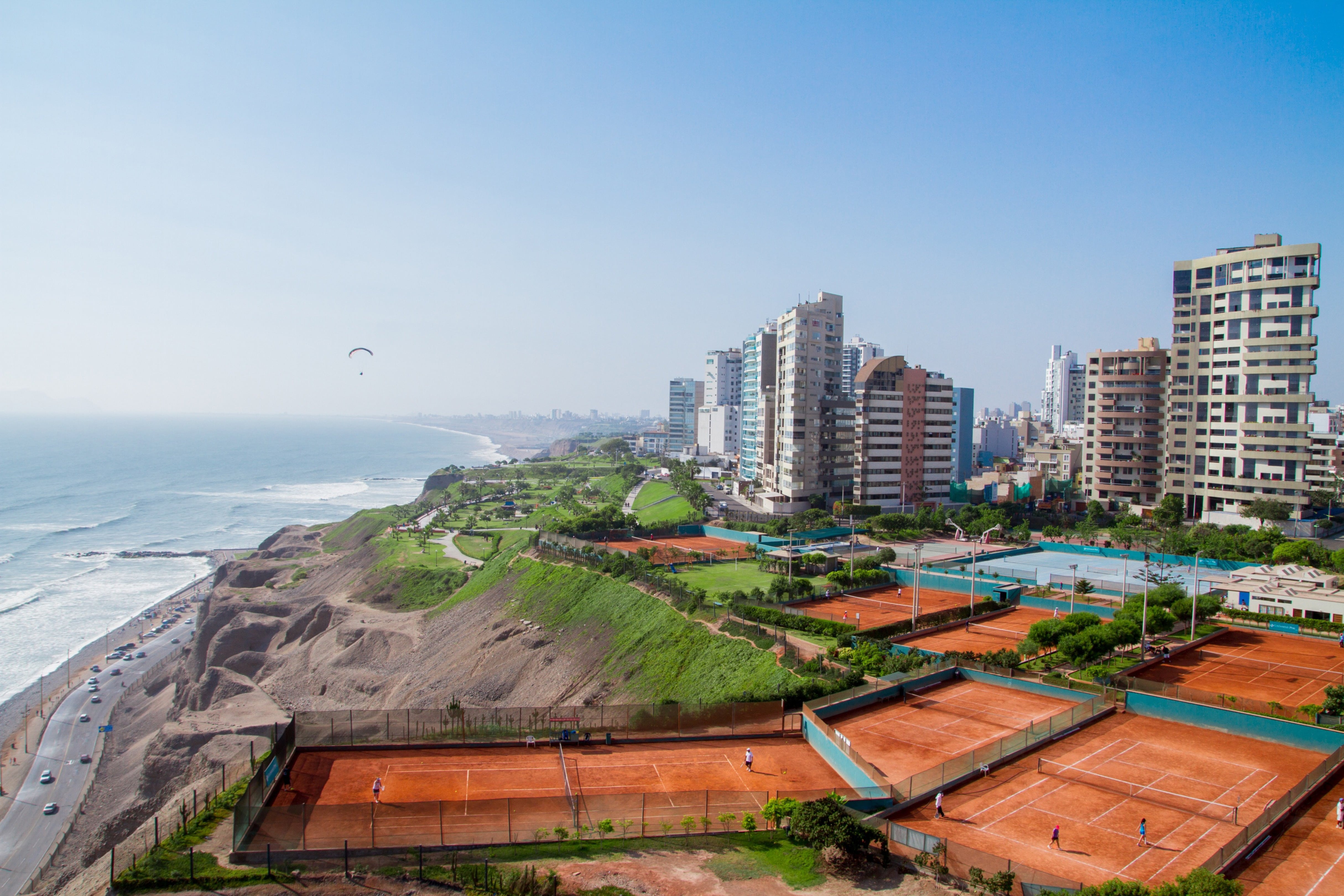 Resa till Lima - Vy över kusten där havet möter stadskanten av Lima i Peru med byggnader, tennisplan och blå himmel