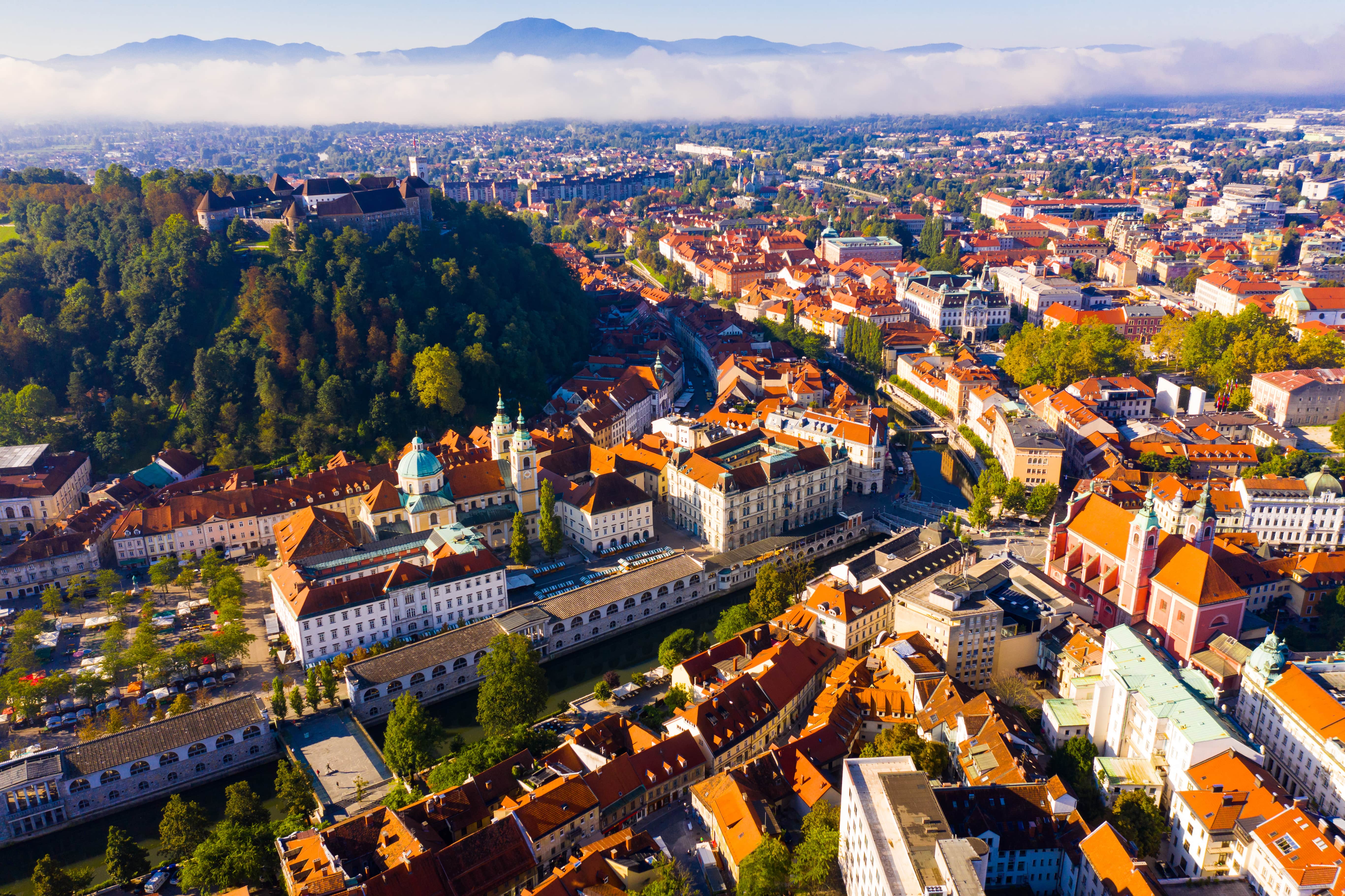 Matkusta Ljubljanaan - Lintuperspektiivi kaupungintalolle, jonka vieressä on vehreää metsää ja taustalla kauniita vuoria ja pilviä